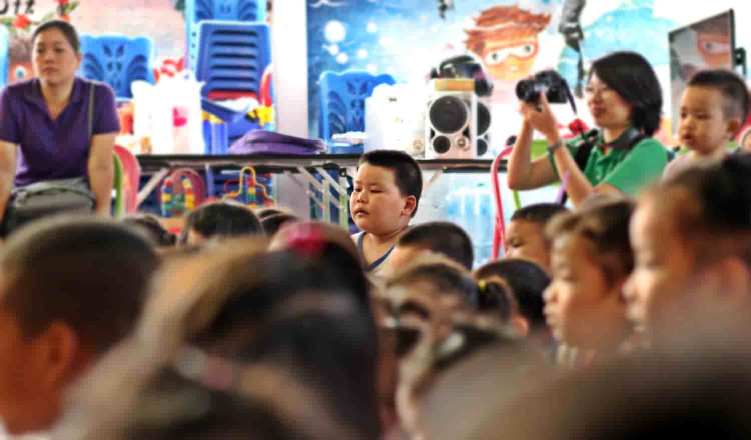 group of children out of focus with one child in focus facing something off screen while an adult behind him takes photos