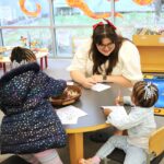 Staff member and two children sitting at a low table. All three are drawing with markers on paper. One of the small children is looking at the staff member while the staff member smiles at them.