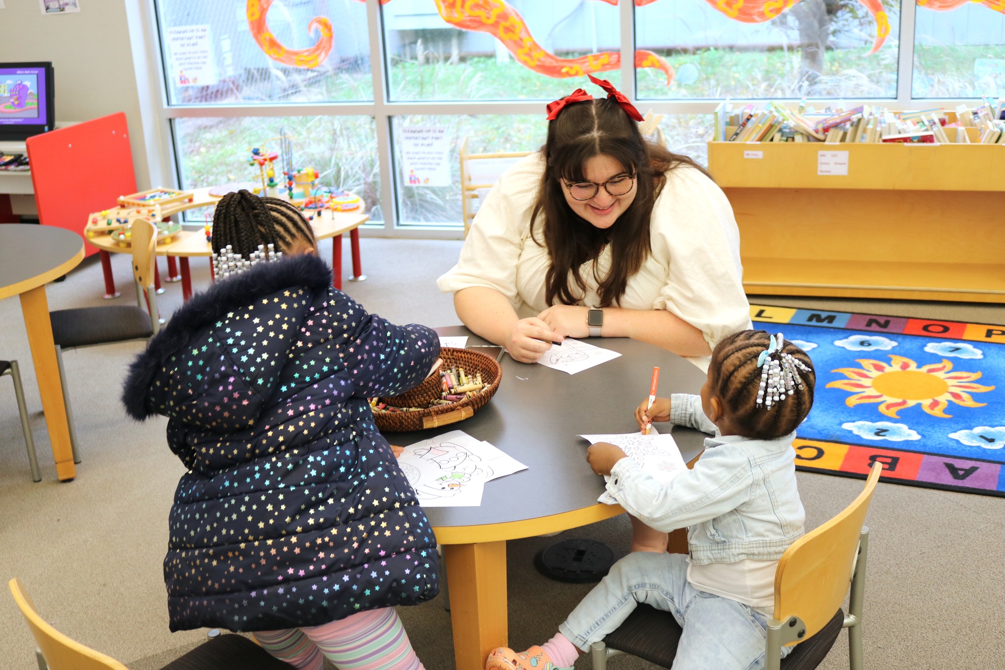 Staff member and two children sitting at a low table. All three are drawing with markers on paper. One of the small children is looking at the staff member while the staff member smiles at them.