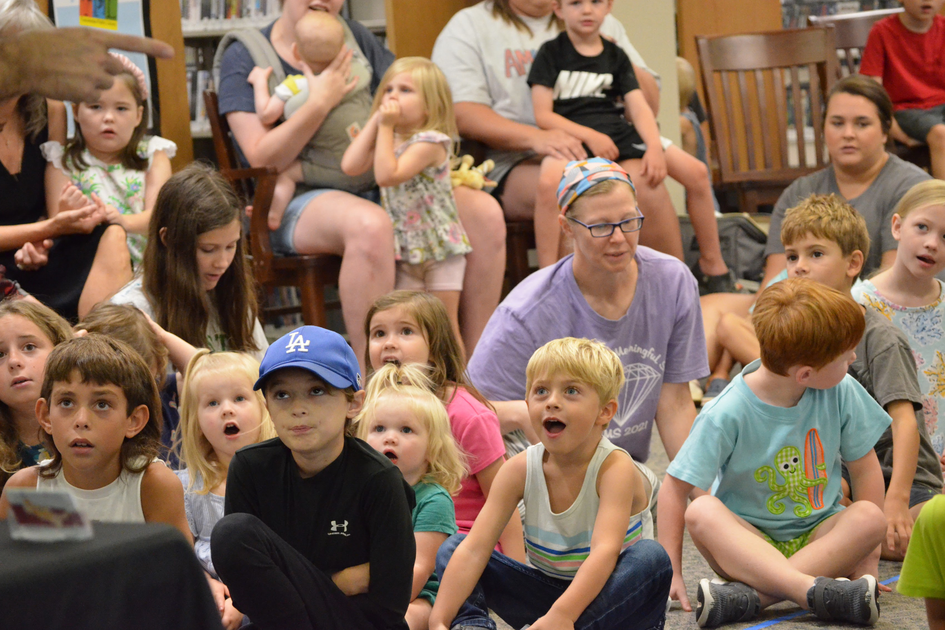 Children sitting on the floor facing and listening to someone out of frame. Adults are sitting behind the students, watching over them.