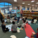 Kids are sitting on the floor surrounding floor mats with squares on them. One of the TPL staff is standing in front of a large television monitor, giving the kids directions. There are square cards with different colors and symbols on them that will be placed on the mats.
