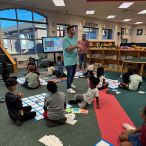 Kids are sitting on the floor surrounding floor mats with squares on them. One of the TPL staff is standing in front of a large television monitor, giving the kids directions. There are square cards with different colors and symbols on them that will be placed on the mats.