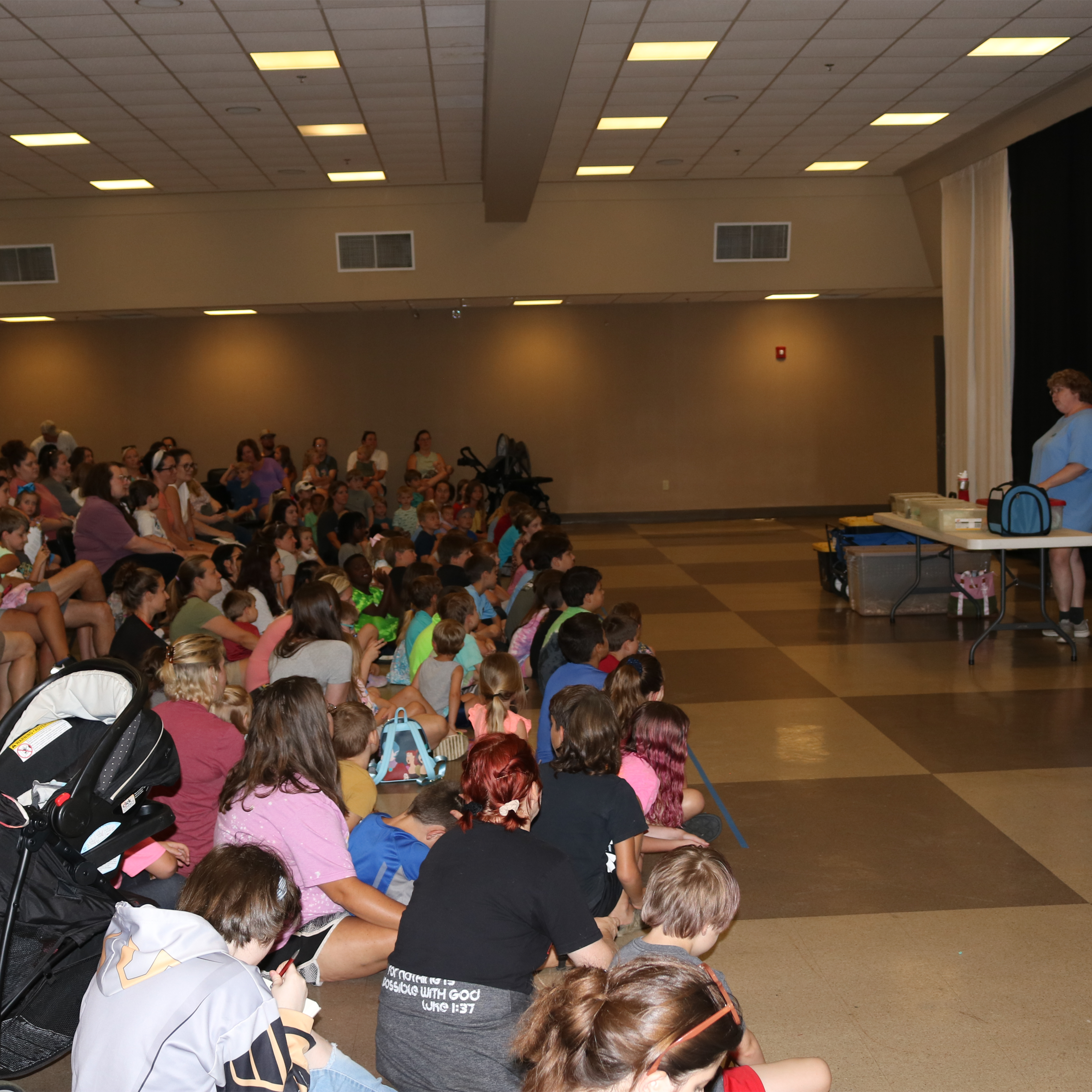 A large group of children and adults of various ages are sitting in rows, facing a staff member who is standing at a table, preparing a demonstration. Children in the front 5-6 rows are seated on the floor while those in the background are seated in chairs.