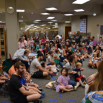 Large group of children and their parents are sitting on the floor inside the main area of the Tuscaloosa Public Library. All of them are watching someone who is out of vision.