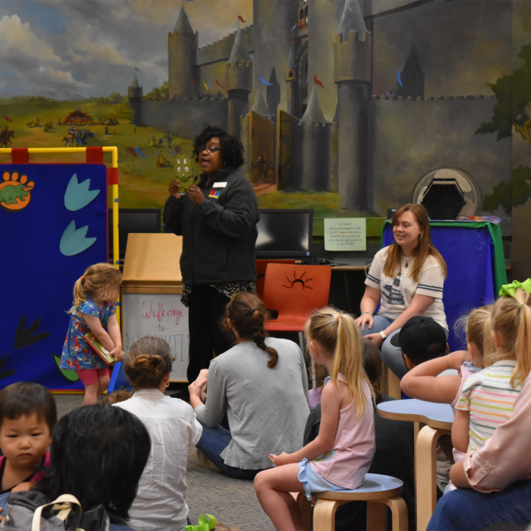 Children are seated on the floor in a room inside the TPL with a backdrop of a castle painted on the wall. One seated and one standing volunteer stand in front of the children. The standing staff member is speaking to the group while everyone watches.