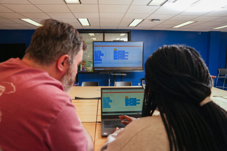 Two individuals looking at a laptop that is also projecting its image on a larger TV screen