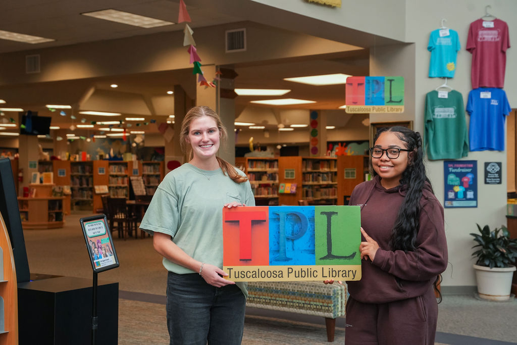 Two women standing in the library entryway holding a sign that says TPL.