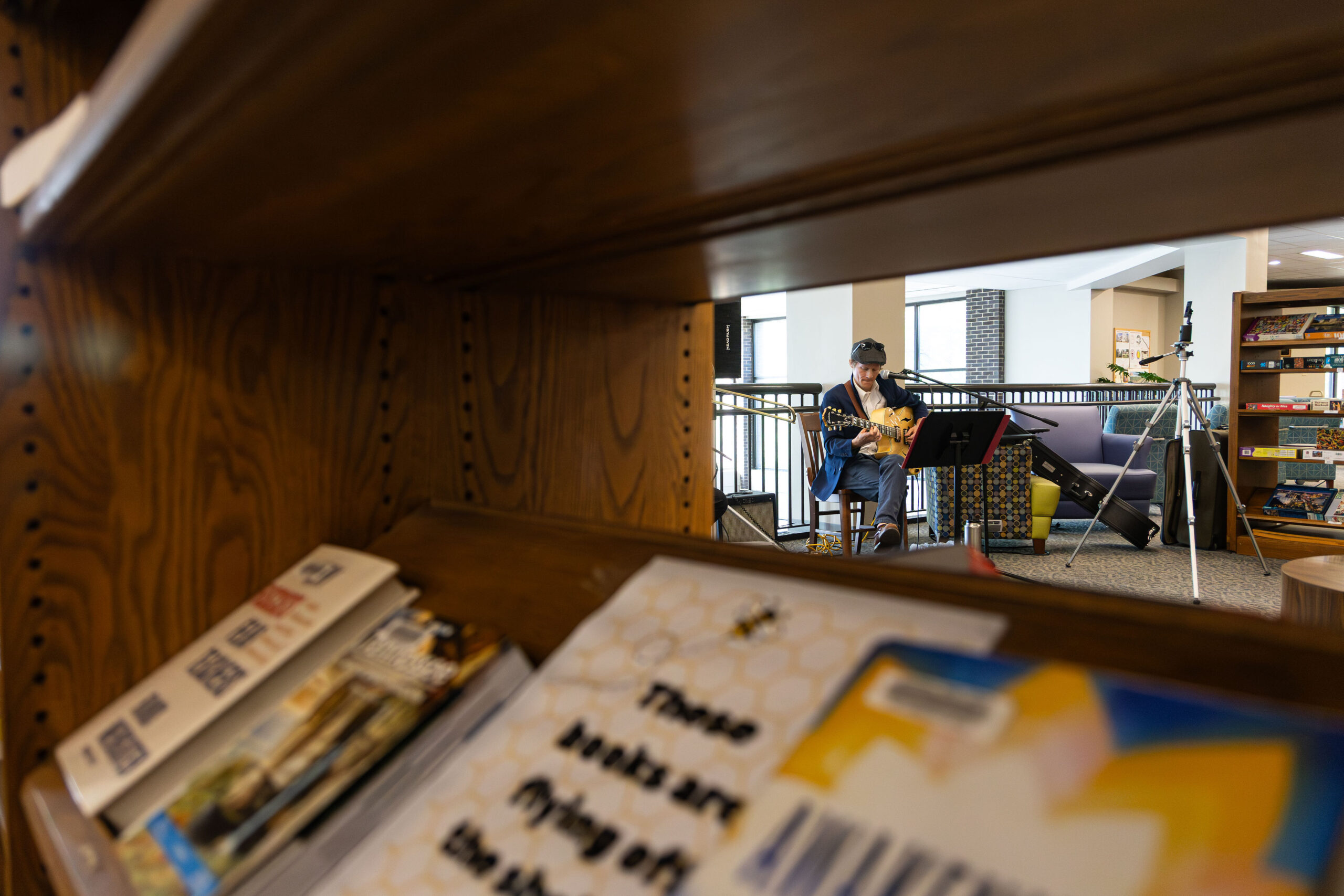 view of a man playing a guitar from the POV of someone peering through the gaps of a bookshelf